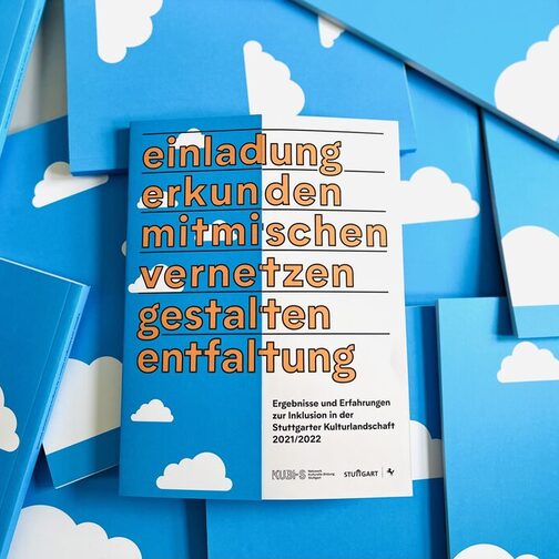 Umgeben von weiteren Broschüren, die auf dem Kopf liegen, liegt die Publikation von KUBI-S mit dem Titel: einladung, erkunden, mitmischen, vernetzen, gestalten, entfaltung" und dem Untertitel: "Ergebnisse und Erfahrungen zur Inklusion in der Stuttgarter Kulturlandschaft 2021/22" sowie den Logos von KUBI-S und der Landeshauptstadt Stuttgart